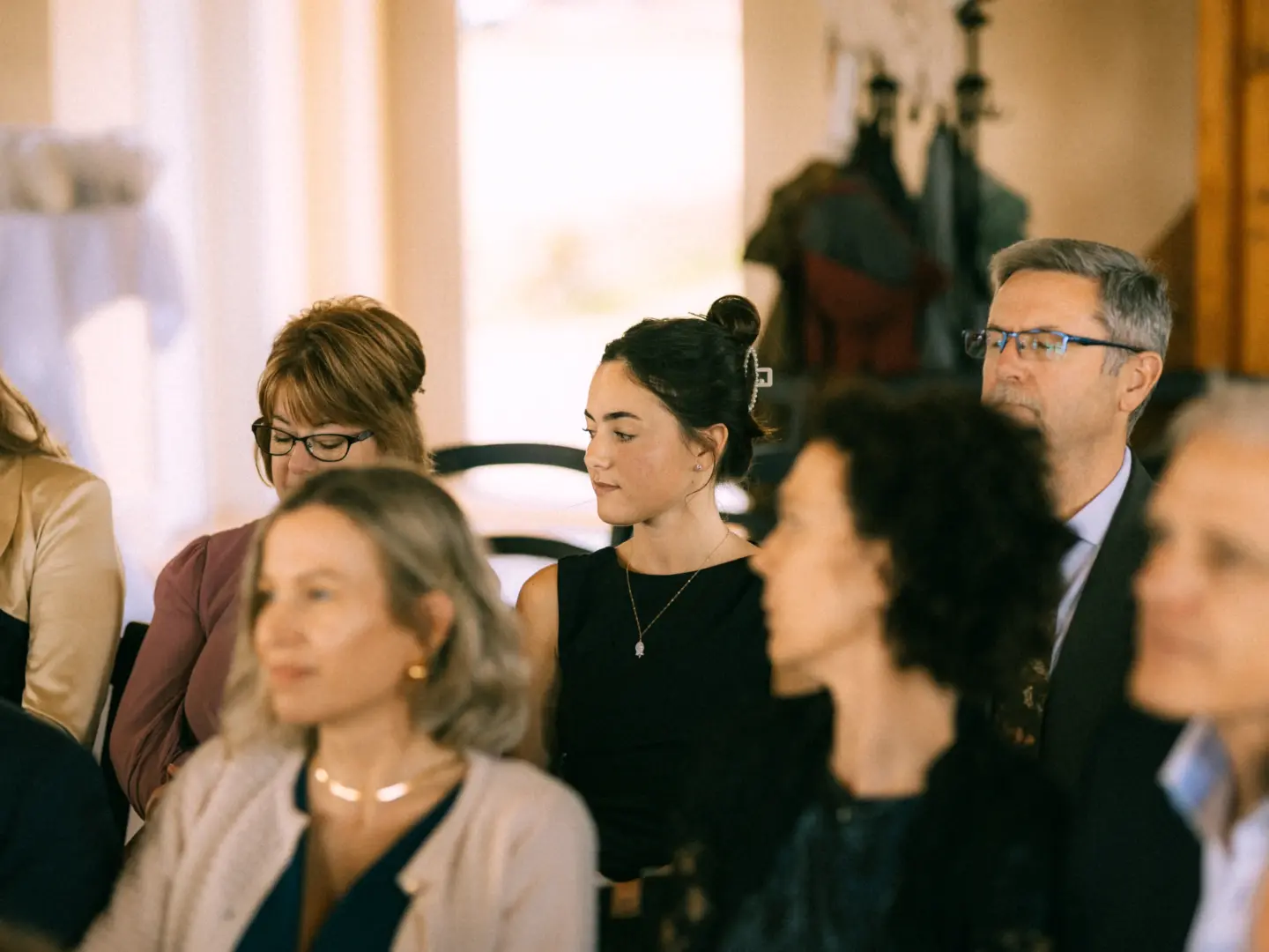 Eine Gruppe von aufmerksam zuhörenden Hochzeitsgästen sitzt in einem hellen Innenraum. Im Fokus steht eine junge Frau mit hochgestecktem Haar, umgeben von weiteren Gästen, bei einer feierlichen Zeremonie, eingefangen von einem Hochzeitsfotograf in Schwerin und MV.