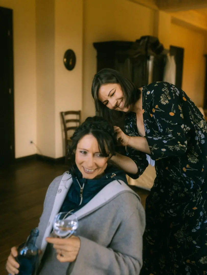 Eine Frau stylt einer anderen lächelnden Frau die Haare bei der Hochzeitsvorbereitung. Die Braut trägt einen grauen Mantel und hält ein Glas Wasser. Professionelle Aufnahmen wie diese entstehen durch einen Hochzeitsfotograf in Schwerin und MV, der die intimen Momente festhält.