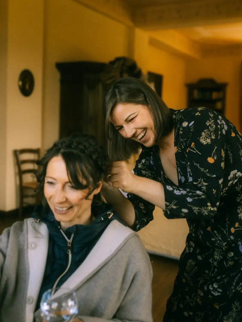 Eine fröhliche Frau mit schulterlangem Haar steckt einer Braut mit geflochtener Frisur eine Blume ins Haar. Die entspannte Vorbereitungsszene fängt die Vorfreude ein – ein besonderer Moment, den ein Hochzeitsfotograf in Schwerin und MV festhält.