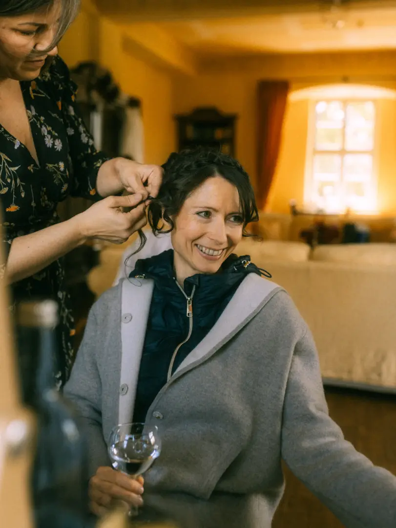 Eine Braut lächelt glücklich, während eine Stylistin ihr die Haare für den großen Tag frisiert. Sie trägt eine graue Jacke über einer dunklen Weste und hält ein Weinglas in der Hand. Die Szene fängt die entspannte Vorbereitung ein, aufgenommen von einem Hochzeitsfotograf in Schwerin und MV.
