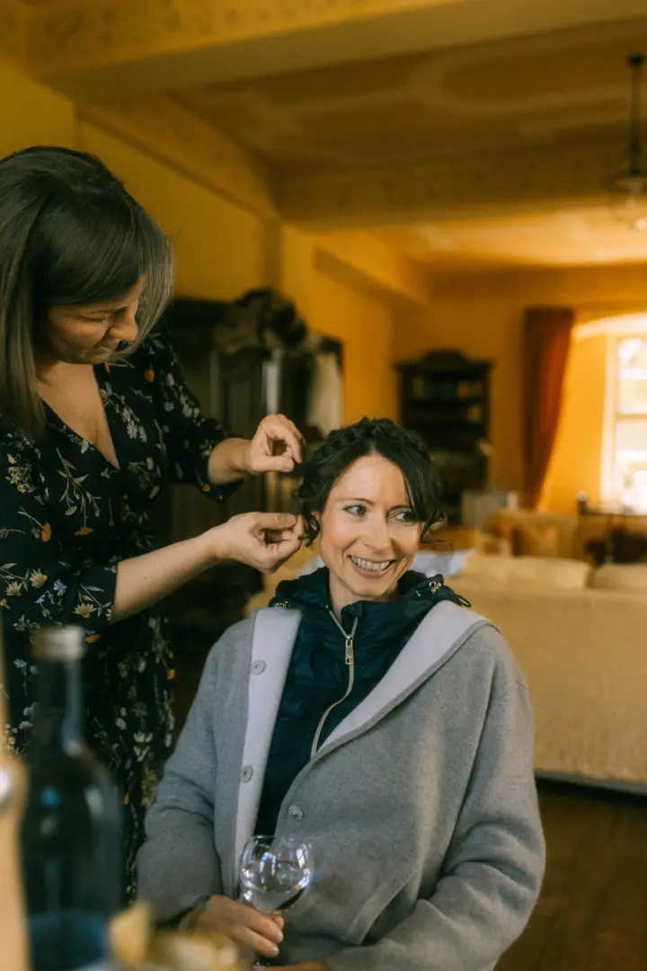 Eine Stylistin steckt einer lächelnden Braut die Haare für die Hochzeit hoch. Die Braut trägt eine graue Strickjacke über einer blauen Jacke und hält ein Weinglas in der Hand. Stimmungsvolle Vorbereitungsszene, festgehalten von einem Hochzeitsfotograf in Schwerin und MV.