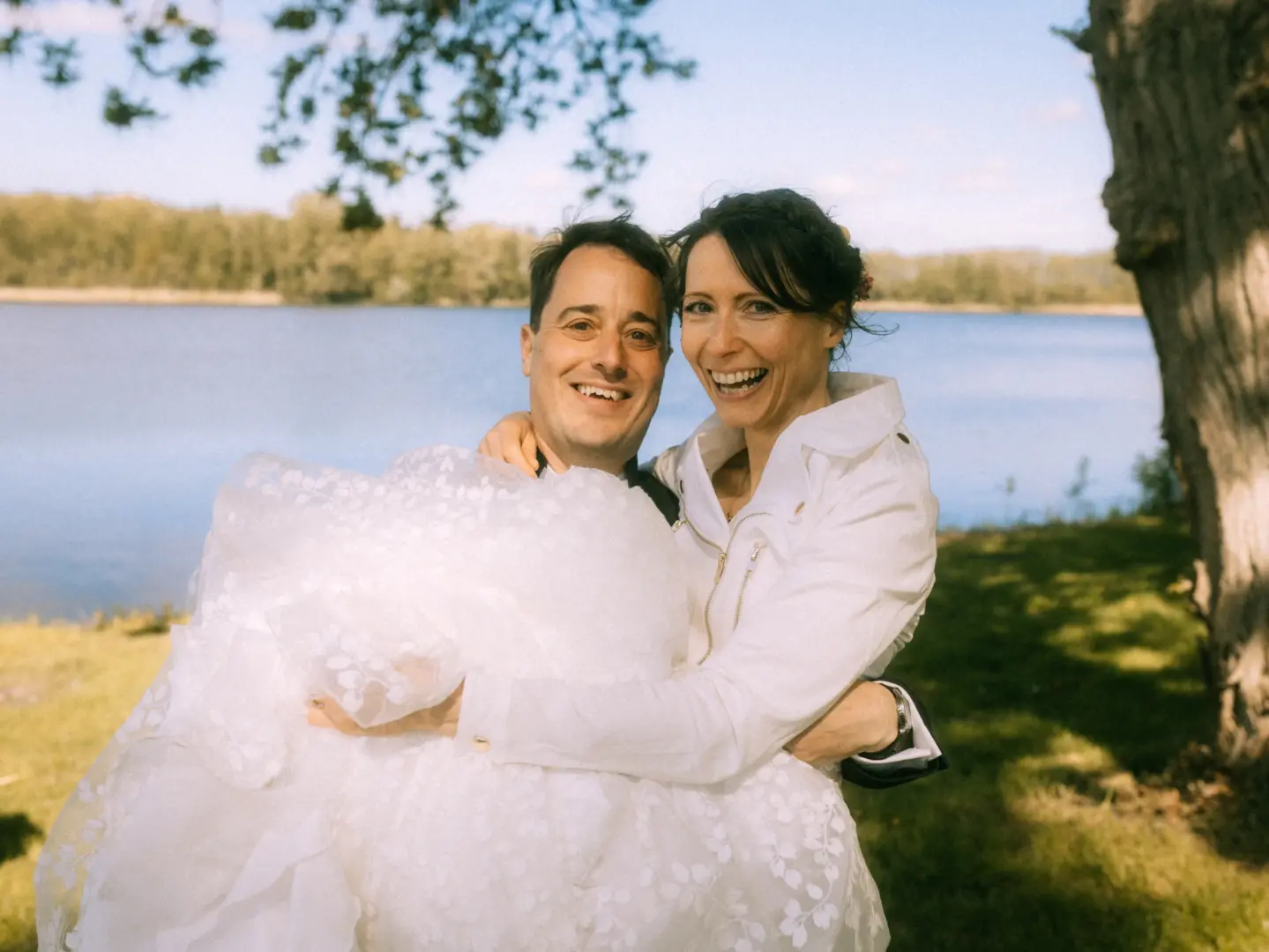 Ein glückliches Brautpaar lacht in die Kamera, während der Bräutigam die Braut in ihrem weißen Spitzenkleid vor einer malerischen See-Kulisse trägt. Ein herzlicher Moment, eingefangen von einem Hochzeitsfotograf in Schwerin und MV.
