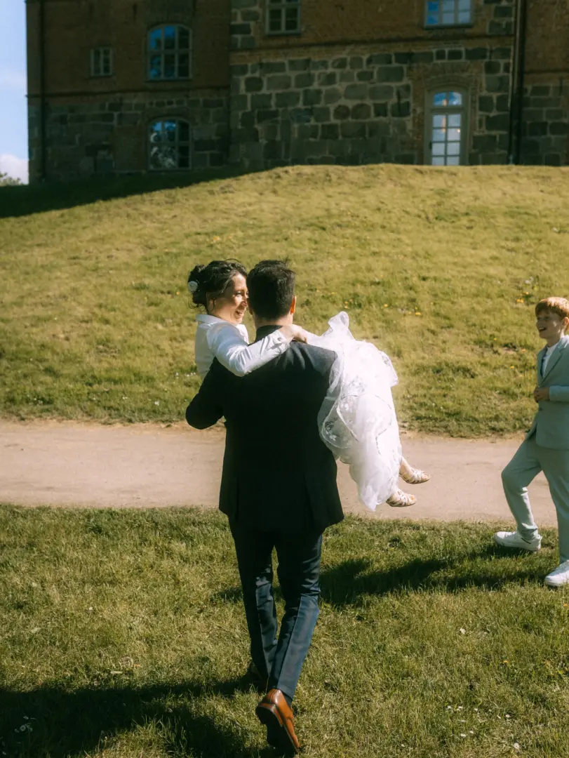 Ein glücklicher Bräutigam trägt seine Braut in einem weißen Hochzeitskleid auf dem Arm, während sie gemeinsam über eine Wiese vor einem historischen Schloss laufen. Ein Junge im Anzug begleitet das Paar. Authentische Momente festgehalten vom Hochzeitsfotograf in Schwerin und MV.