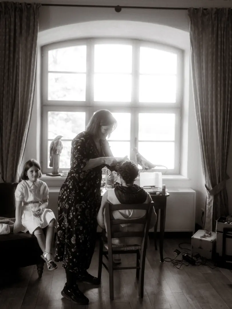 Eine Stylistin frisiert eine Braut vor einem hellen Fenster in einem eleganten Raum, während ein junges Mädchen geduldig daneben sitzt. Diese intime Vorbereitungsszene wurde von einem Hochzeitsfotograf in Schwerin und MV festgehalten.