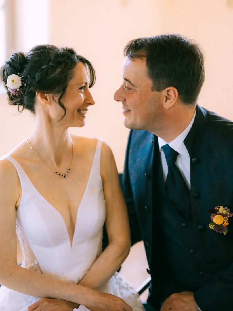Ein glückliches Brautpaar blickt sich bei der Trauung liebevoll an. Die Braut trägt ein weißes Brautkleid mit tiefem Ausschnitt und Blumen im Haar, der Bräutigam einen eleganten dunklen Anzug mit Ansteckblume. Eine emotionale Aufnahme vom Lovestory Fotograf in Schwerin und MV.