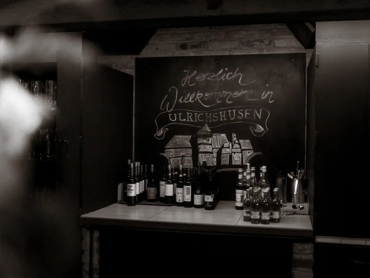 Eine festliche Bar in Ulrichshusen mit einer handgeschriebenen Kreidetafel „Herzlich Willkommen in Ulrichshusen“ über einer Auswahl an Weinflaschen. Das stimmungsvolle Ambiente eignet sich perfekt für Feiern, festgehalten von einem Hochzeitsfotograf Müritz und MV.