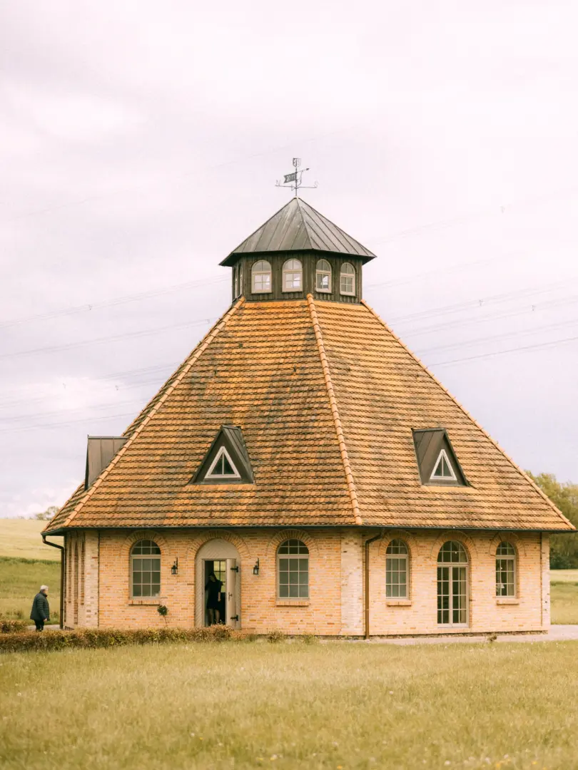 Ein elegantes, achteckiges Backsteingebäude mit einem markanten Zeltdach und einer kleinen Laterne an der Spitze, umgeben von einer weiten Wiesenlandschaft. Eine Person tritt durch den offenen Eingang in das Gebäude, das als stilvolle Location für einen Hochzeitsfotograf in Schwerin und MV dient.