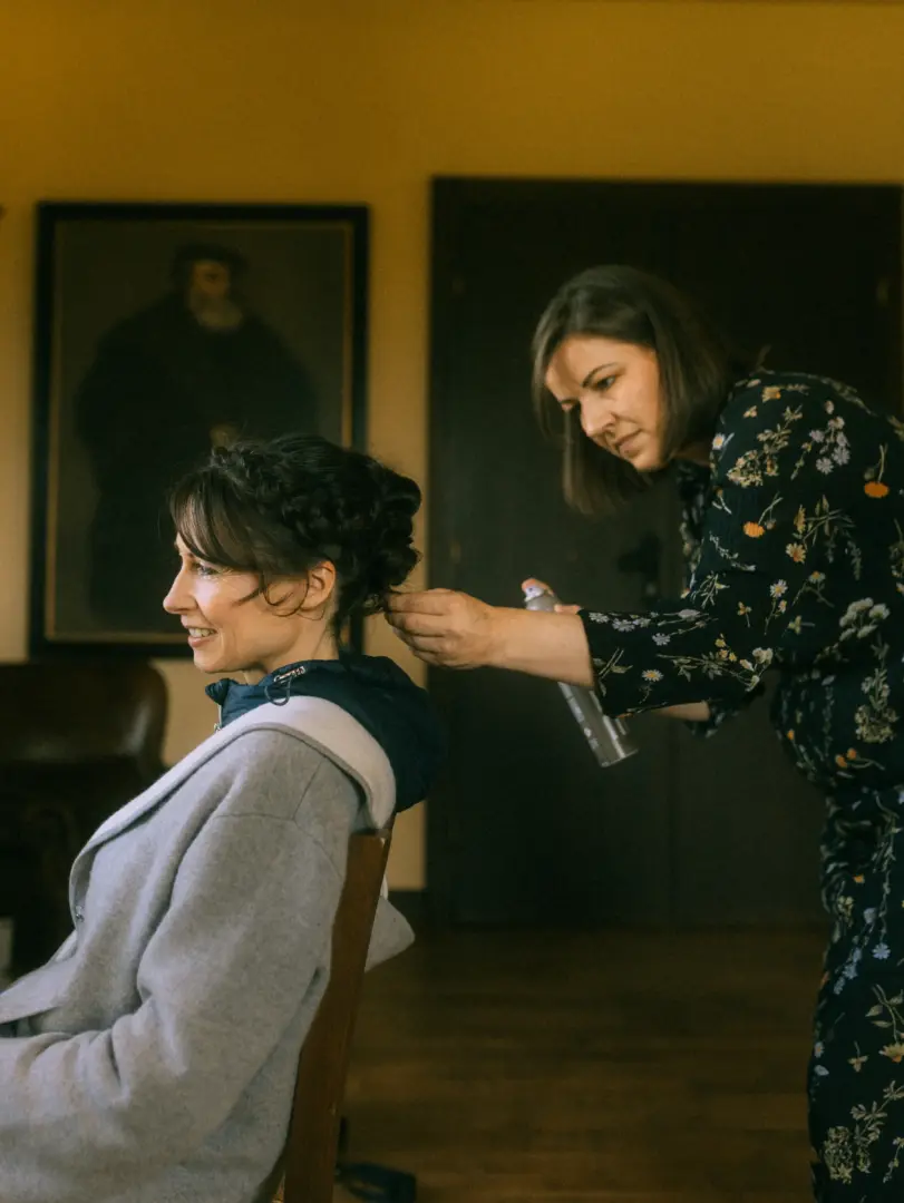 Eine Braut mit kunstvoll geflochtener Hochsteckfrisur sitzt entspannt auf einem Stuhl, während eine Stylistin ihr Haar mit Haarspray fixiert. Die Szene fängt die intime Vorbereitung durch einen professionellen Hochzeitsfotograf in Schwerin und MV ein.