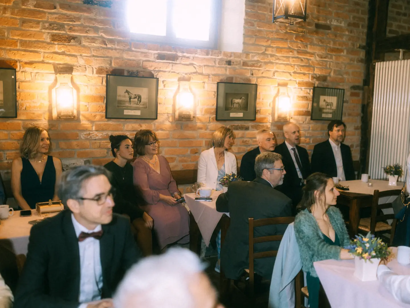 Gäste sitzen an festlich gedeckten Tischen in einem rustikalen Raum mit Ziegelwänden, der von einem Hochzeitsfotograf in Schwerin und MV fotografiert wurde. Die Szene zeigt eine elegante Hochzeitsgesellschaft in entspannter, gehobener Atmosphäre bei einer Feier.