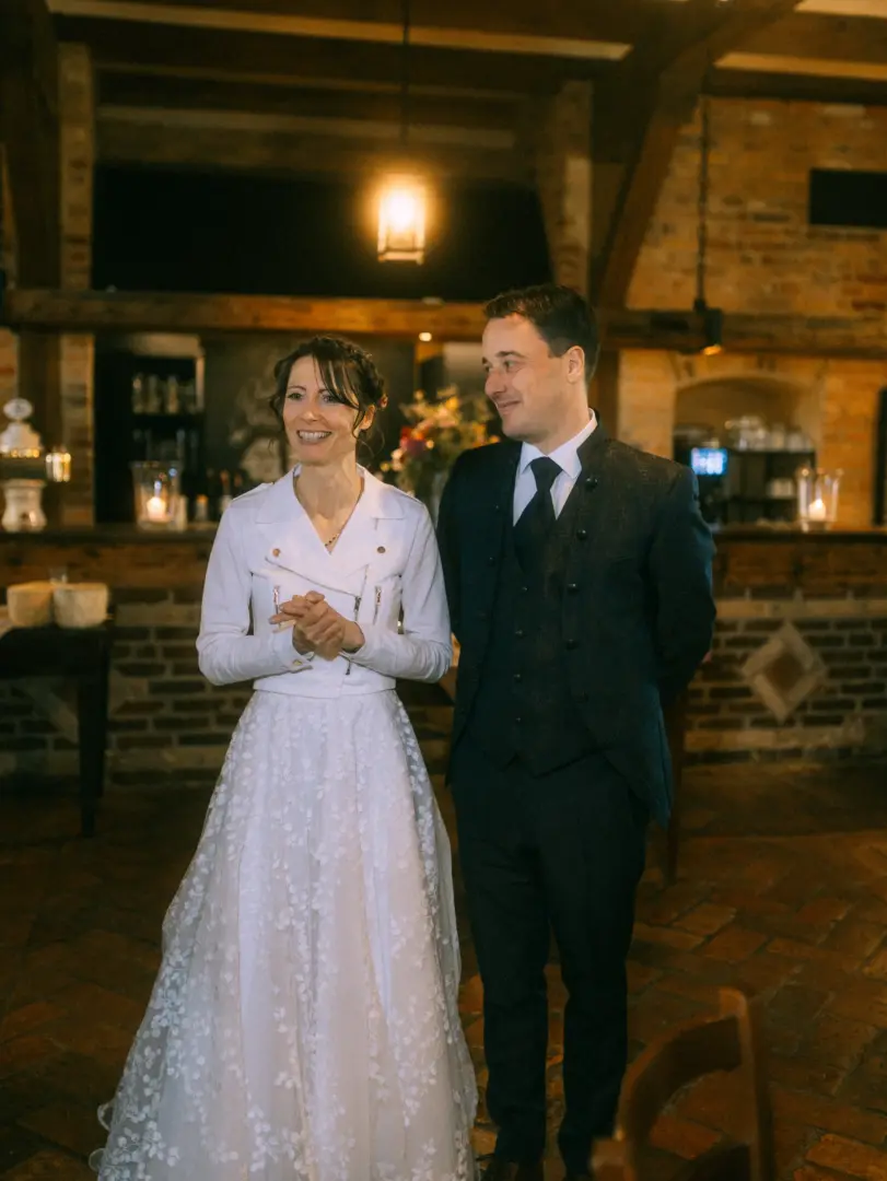Ein glückliches Brautpaar steht in einer rustikalen Location mit Backsteinwänden. Die Braut trägt ein weißes Kleid mit floralem Muster und eine Bikerjacke, der Bräutigam einen eleganten dunklen Anzug. Eine stimmungsvolle Aufnahme von einem Hochzeitsfotograf in Schwerin und MV.