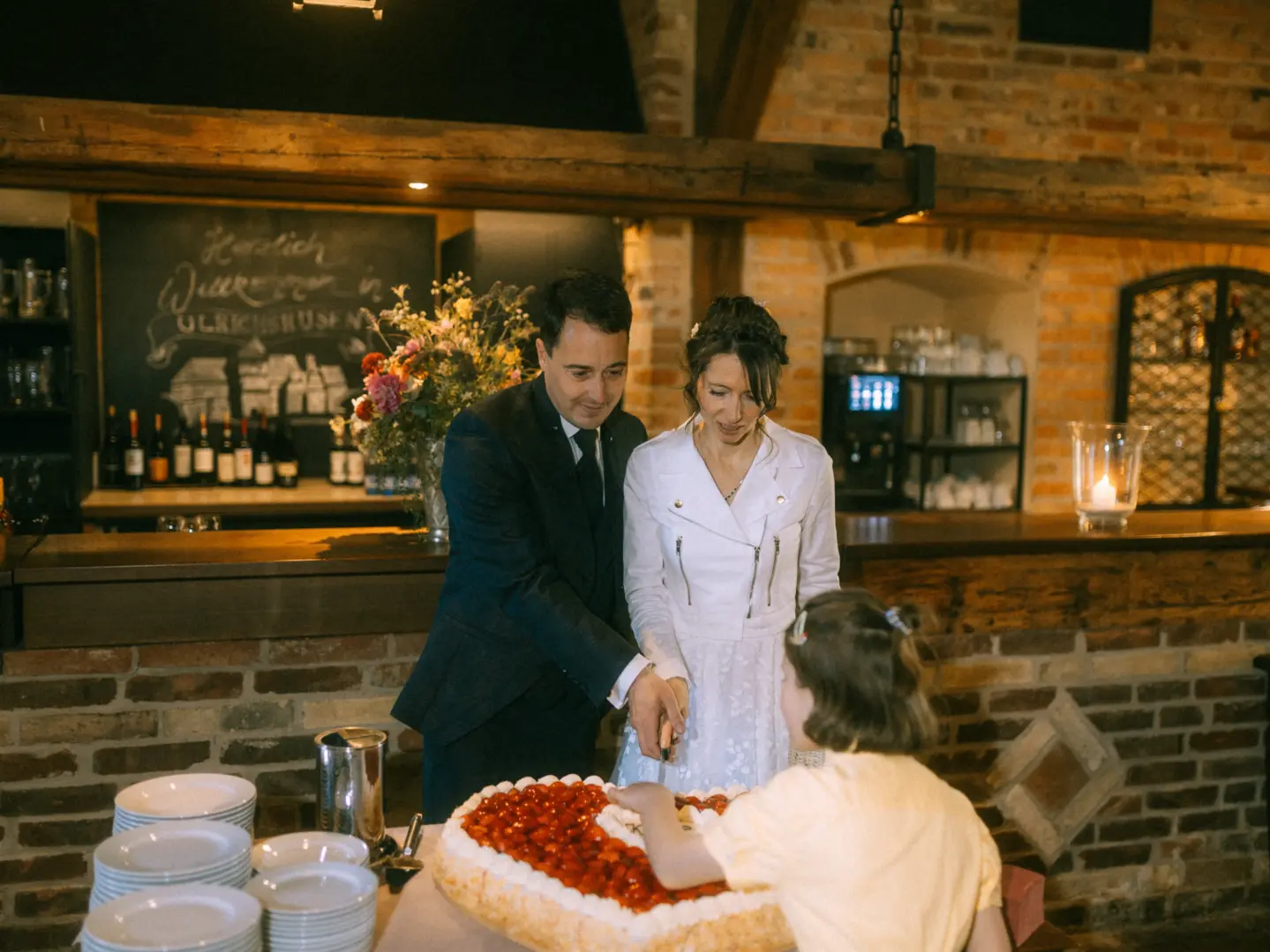 Ein Brautpaar schneidet gemeinsam mit einem Kind eine herzförmige Erdbeertorte in einer rustikalen Location mit Backsteinwänden an. Im Hintergrund steht eine Tafel mit der Aufschrift „Herzlich Willkommen in Ulrichshusen“. Hochzeitsfotograf in Schwerin und MV.