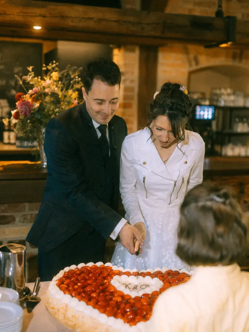 Ein Brautpaar schneidet gemeinsam eine herzförmige Erdbeertorte mit der Inschrift „K&C“ an. Die Braut trägt eine weiße Jacke, der Bräutigam einen schwarzen Anzug. Eine stimmungsvolle Aufnahme vom Hochzeitsfotograf in Schwerin und MV in einer rustikalen Location.
