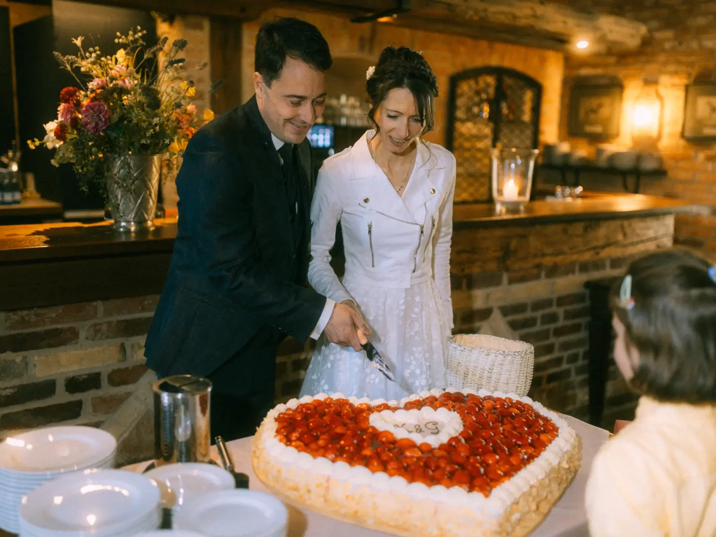Ein Brautpaar schneidet gemeinsam eine herzförmige Hochzeitstorte mit Erdbeeren und der Inschrift „K & G“ an. Die Braut trägt eine weiße Bikerjacke über ihrem Kleid, während der Bräutigam im dunklen Anzug strahlt. Hochzeitsfotograf in Schwerin und MV festgehalten in rustikalem Ambiente.