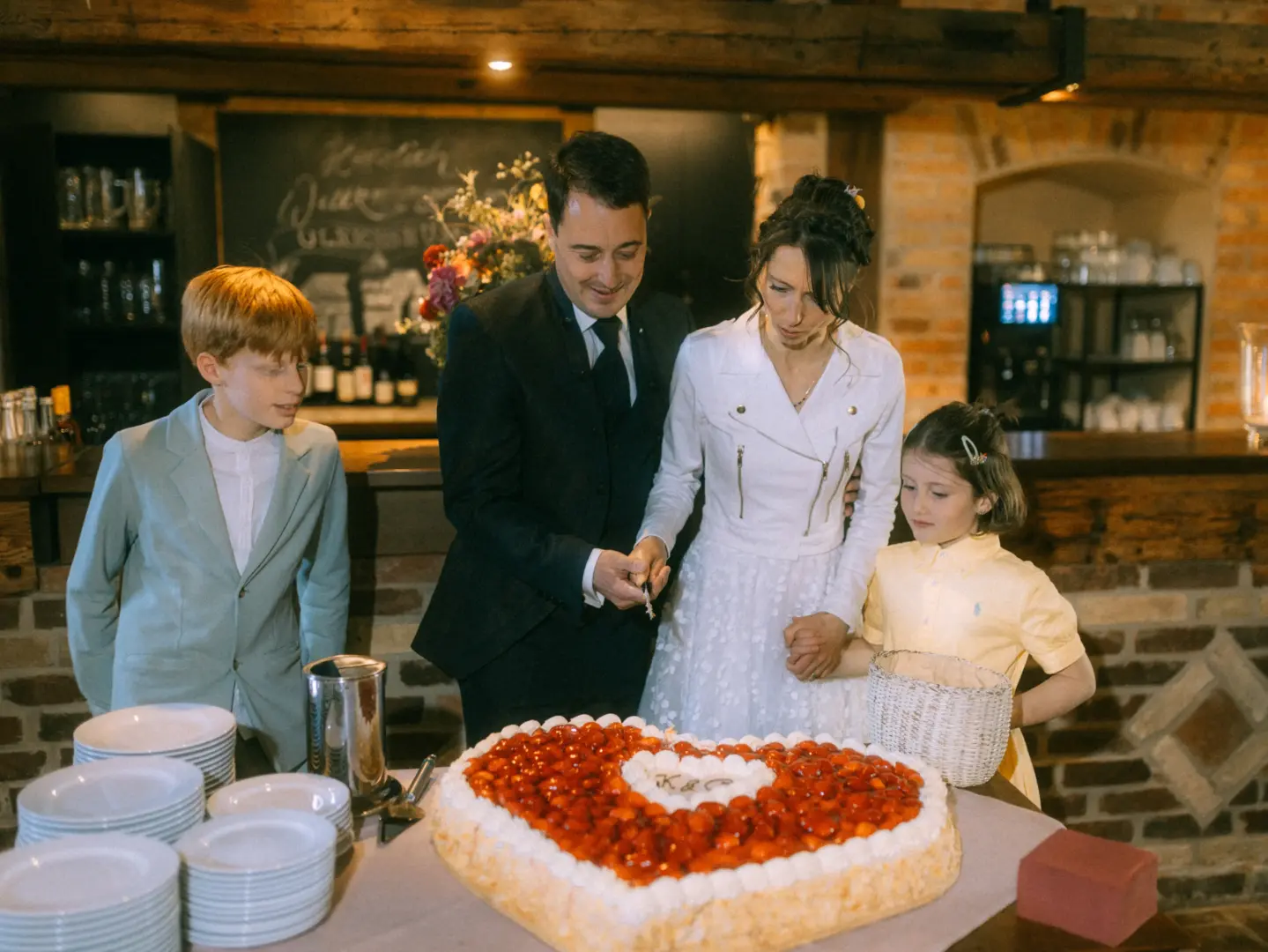 Ein Brautpaar schneidet gemeinsam mit zwei Kindern eine herzförmige Erdbeertorte an. Die Szene ist in einem rustikalen Restaurant mit Backsteinwänden aufgenommen. Professionelle Hochzeitsfotografie von einem Hochzeitsfotograf in Schwerin und MV.