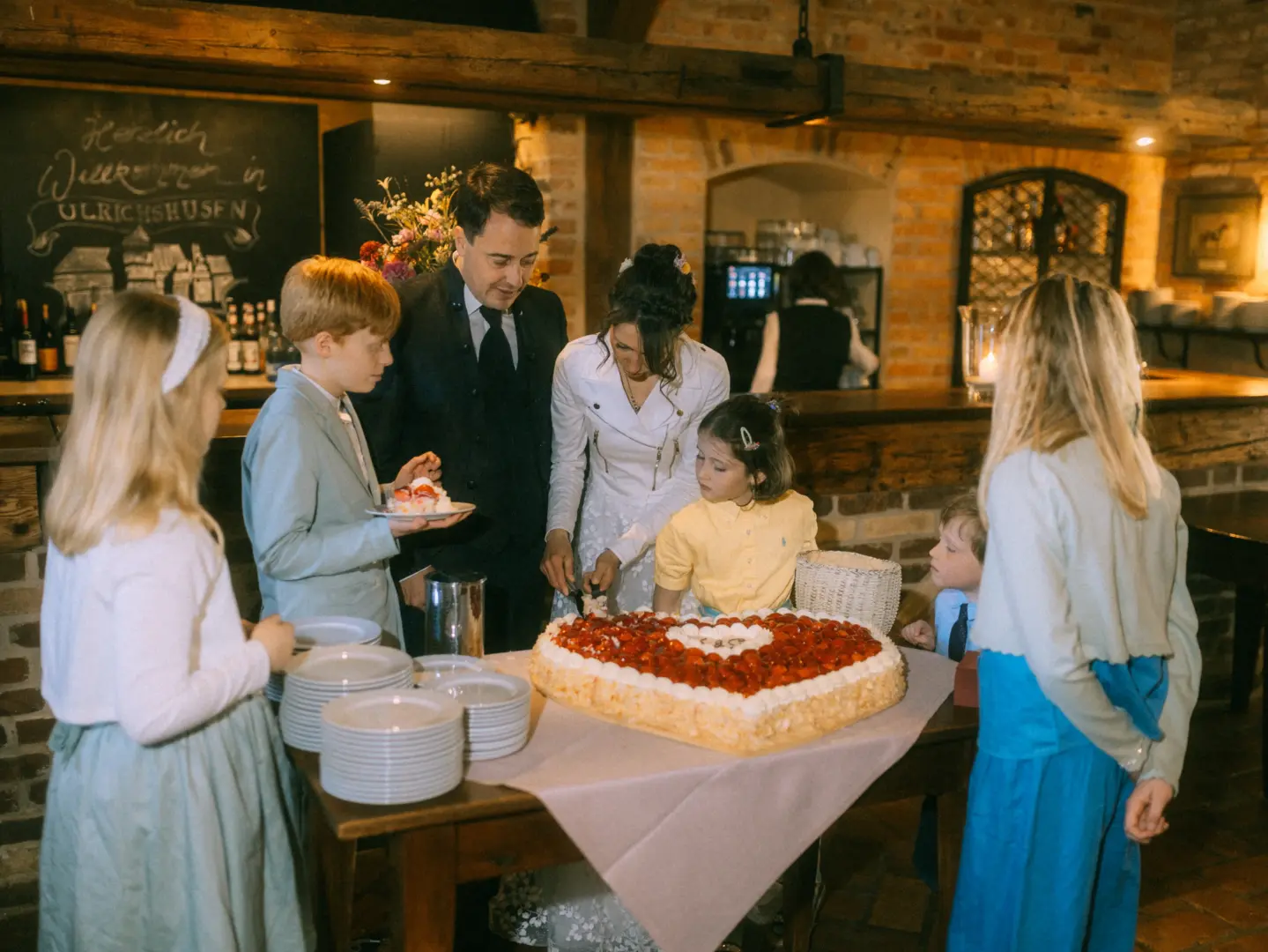 Ein Brautpaar schneidet gemeinsam eine herzförmige Erdbeertorte an, umringt von Kindern in einer rustikalen Location mit einer Tafel „Herzlich Willkommen in Ulrichshusen“. Stimmungsvolle Aufnahme vom Hochzeitsfotograf Müritz und MV.