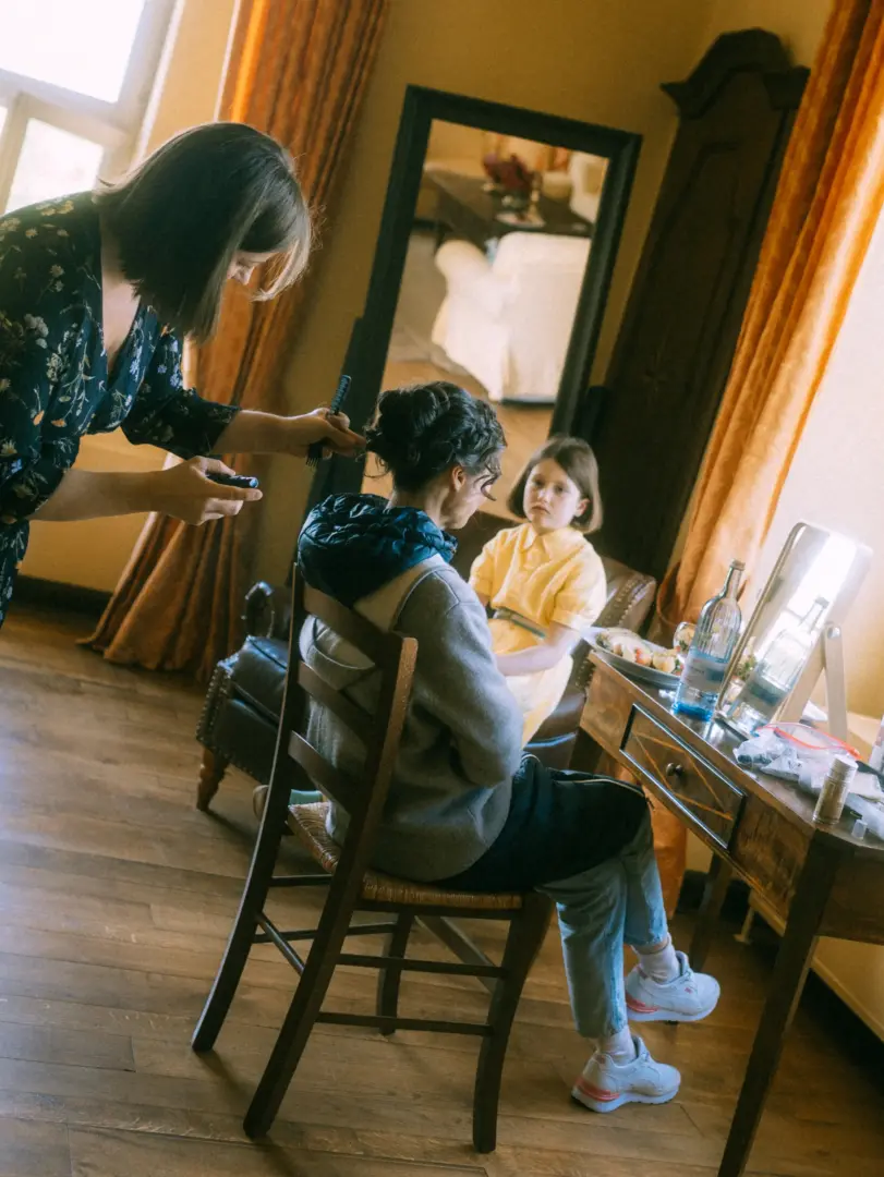 Eine Stylistin frisiert eine Braut in legerer Kleidung, während ein kleines Mädchen in einem gelben Kleid im Spiegel beobachtet wird. Diese intime Vorbereitungsszene wurde von einem Hochzeitsfotograf in Schwerin und MV festgehalten.