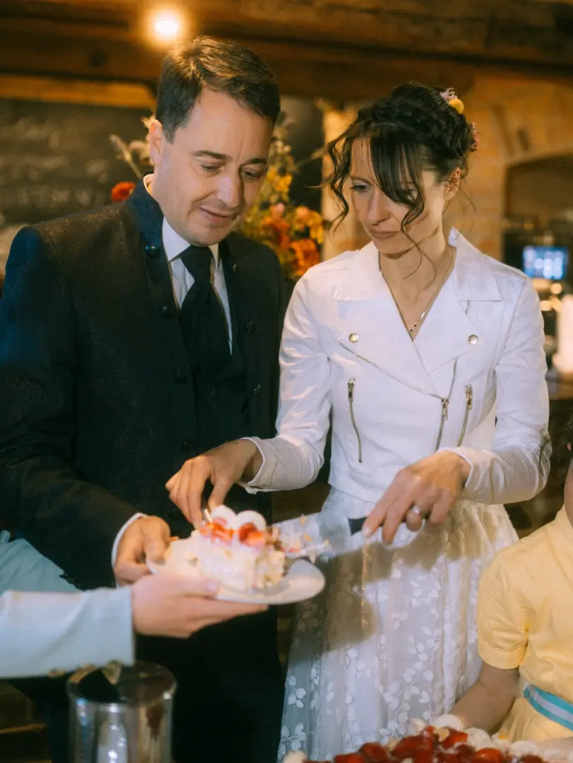 Ein Brautpaar schneidet gemeinsam die Hochzeitstorte an: Die Braut trägt eine weiße Lederjacke über ihrem Hochzeitskleid, der Bräutigam einen eleganten schwarzen Anzug. Ein Lovestory Fotograf in Schwerin und MV hat diesen intimen Moment der Feierlichkeit stimmungsvoll festgehalten.