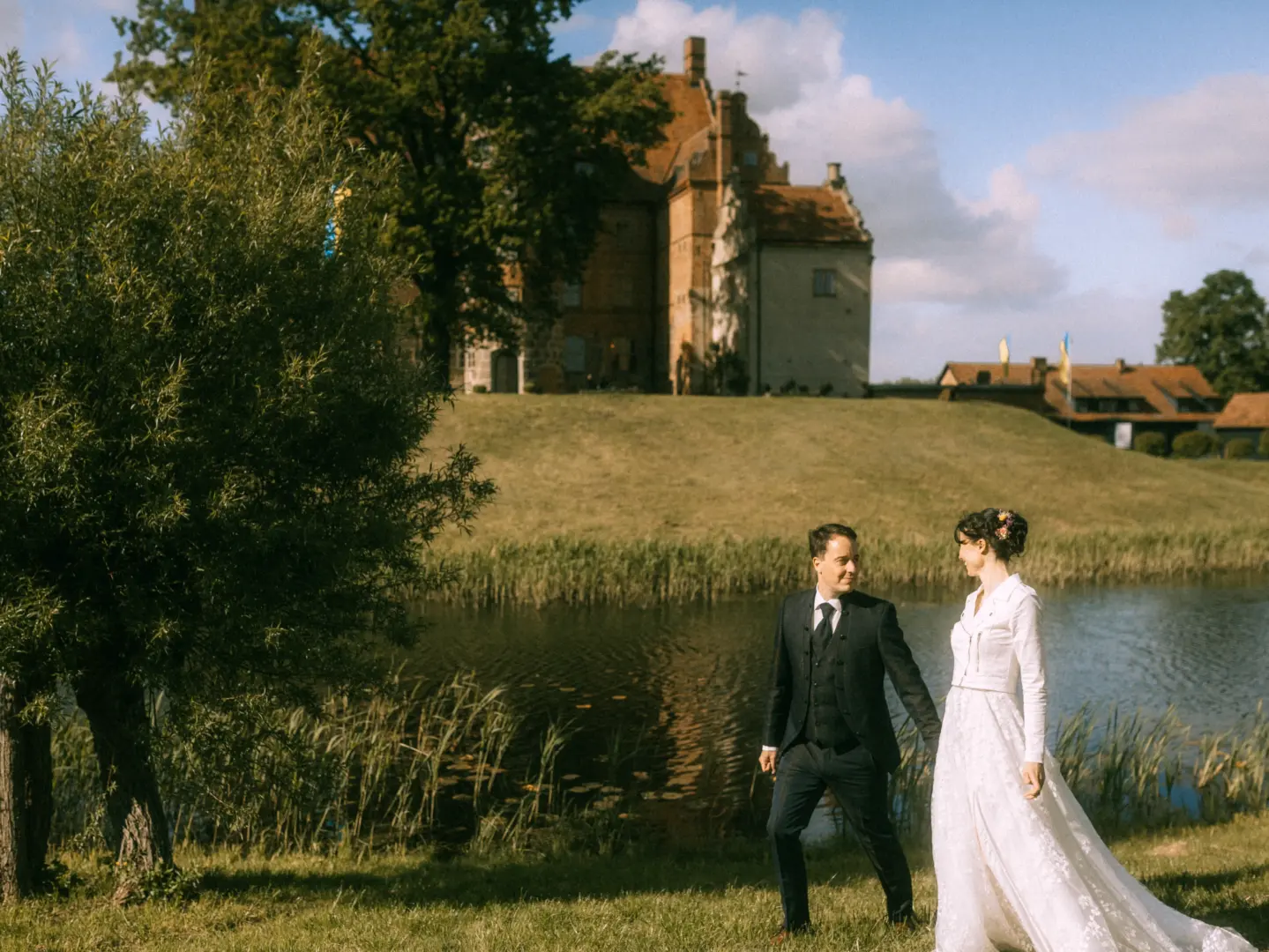 Ein Brautpaar spaziert händchenhaltend vor einem historischen Schloss mit Wassergraben. Die Braut trägt ein weißes Spitzenkleid mit Jäckchen, der Bräutigam einen eleganten Anzug. Romantische Hochzeitsfotograf in Schwerin und MV Aufnahme in einer malerischen Parklandschaft bei Sonnenschein.