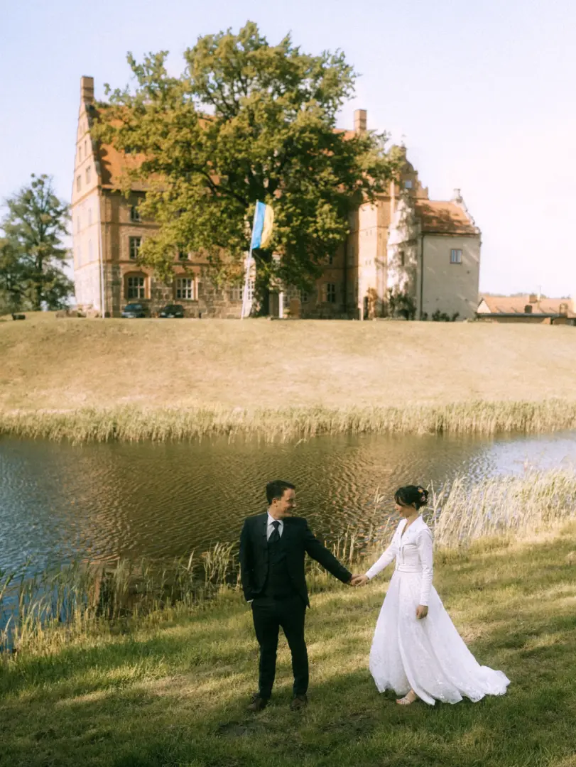 Ein Brautpaar hält sich an den Händen vor einem historischen Schloss mit Wassergraben. Die Braut trägt ein weißes Kleid mit Jacke, der Bräutigam einen dunklen Anzug. Eine ukrainische Flagge weht im Hintergrund. Hochzeitsfotograf in Schwerin und MV.