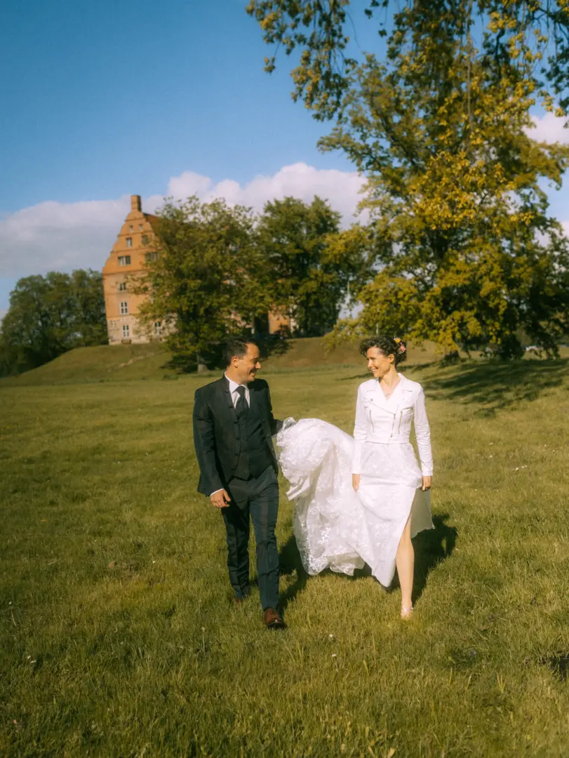 Ein glückliches Brautpaar spaziert lachend über eine sonnige Wiese vor einem historischen Schloss, während der Bräutigam das Kleid der Braut hält. Eine romantische Momentaufnahme von einem Lovestory Fotograf in Schwerin und MV, die eine unbeschwerte Hochzeitsatmosphäre einfängt.