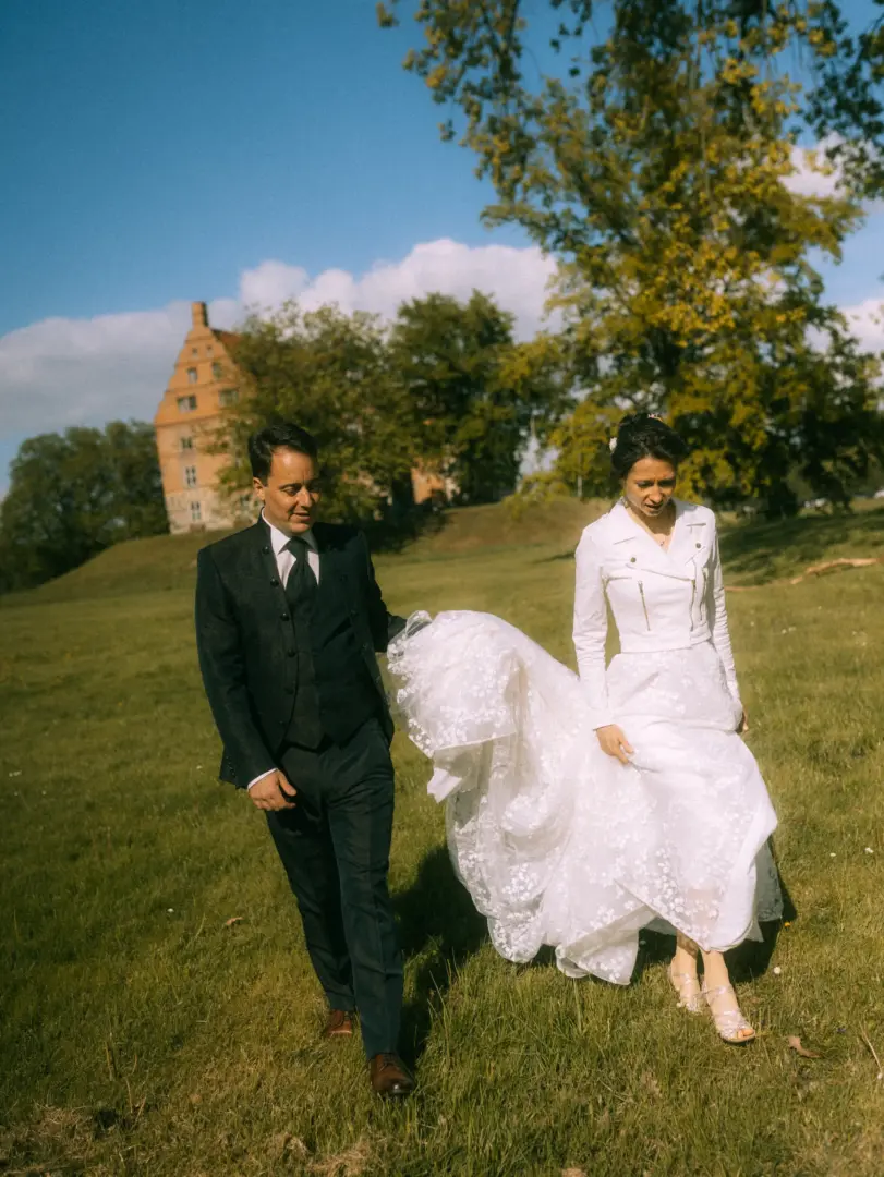 Ein Brautpaar spaziert glücklich über eine sonnige Wiese vor einem historischen Schloss. Die Braut trägt eine weiße Lederjacke über ihrem Hochzeitskleid, der Bräutigam einen eleganten dunklen Anzug. Hochzeitsfotograf in Schwerin und MV.