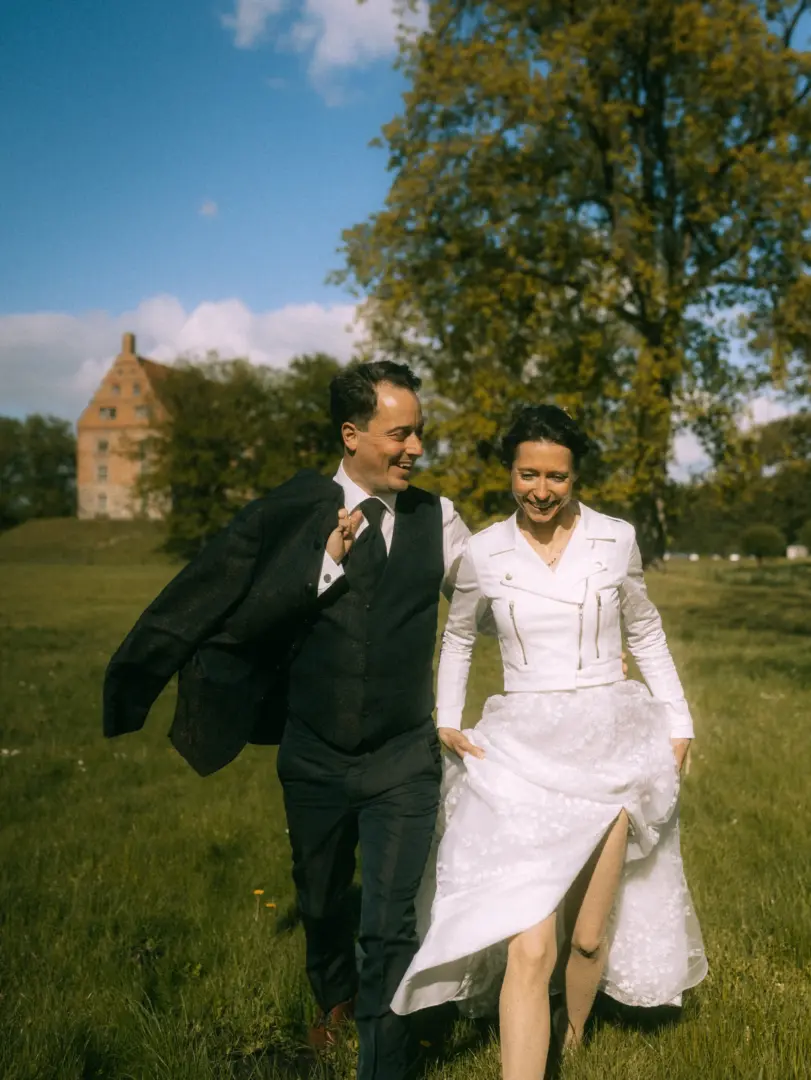 Ein glückliches Brautpaar läuft lachend über eine Wiese vor einem historischen Schloss. Die Braut trägt eine weiße Lederjacke zum Brautkleid, der Bräutigam einen eleganten Anzug. Authentische Hochzeitsfotografie als Hochzeitsfotograf in Schwerin und MV.