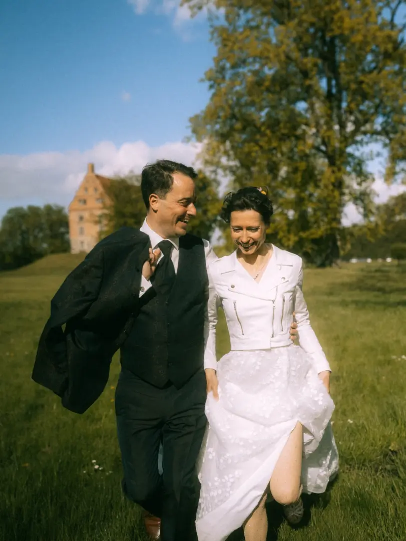 Ein glückliches Brautpaar läuft lachend über eine sonnige Wiese vor einem historischen Schloss. Die Braut trägt eine weiße Bikerjacke zum Hochzeitskleid, der Bräutigam einen eleganten Anzug. Authentische Hochzeitsfotograf in Schwerin und MV Aufnahme voller Lebensfreude.