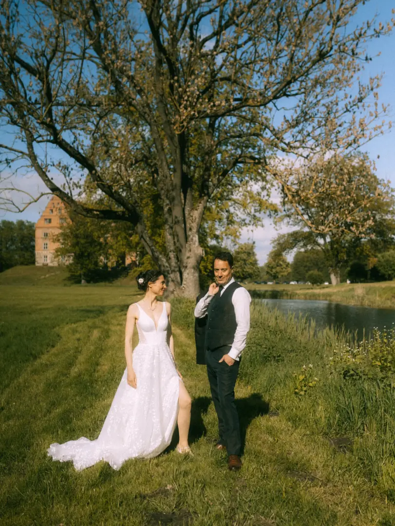 Ein Brautpaar posiert an einem sonnigen Tag vor einem historischen Schloss und einem großen Baum. Die Braut trägt ein weißes Spitzenkleid mit Beinschlitz, der Bräutigam einen eleganten Anzug. Lovestory Fotograf in Schwerin und MV für unvergessliche Hochzeitsmomente im Freien.