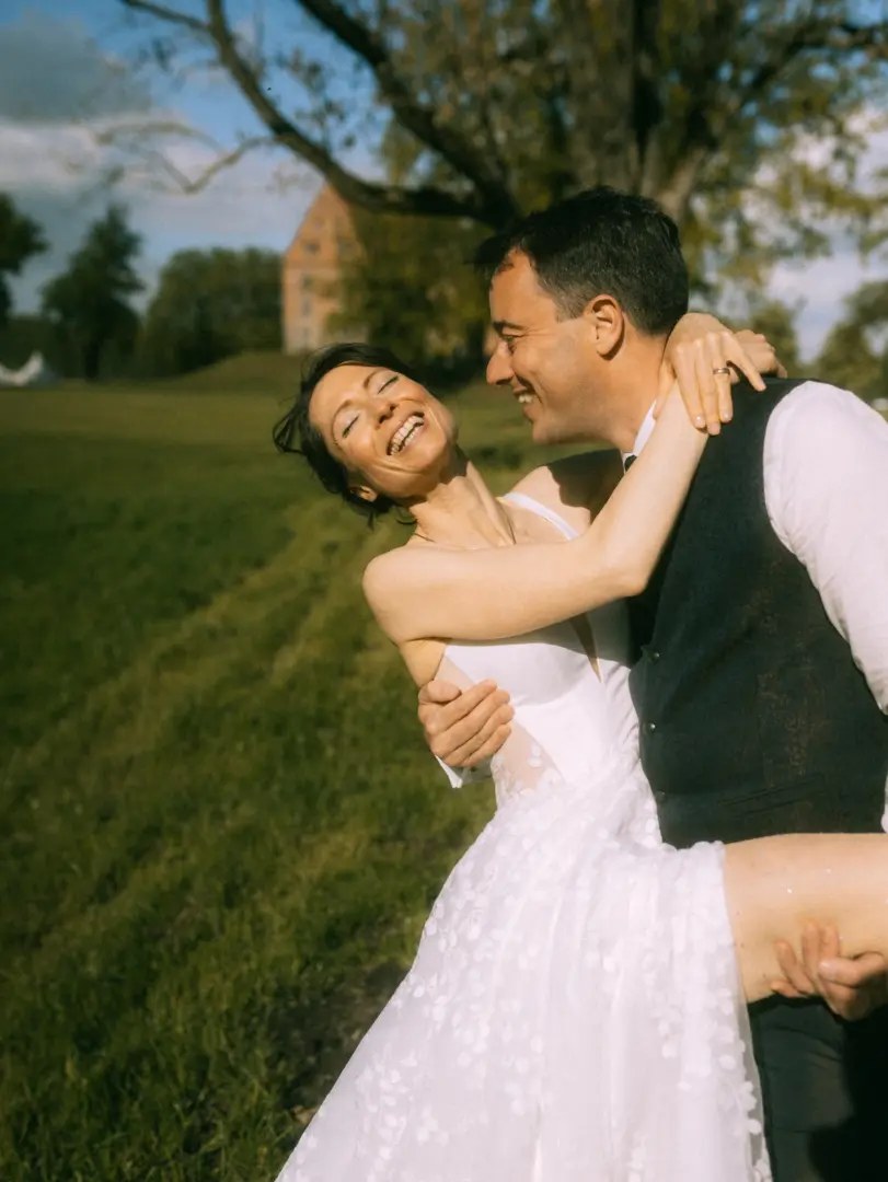 Ein glückliches Brautpaar bei einem romantischen Moment im Freien: Der Bräutigam hebt die lachende Braut in ihrem weißen Hochzeitskleid hoch, während sie sich eng umarmen. Im Hintergrund erstreckt sich eine grüne Wiese und ein Schloss. Lovestory Fotograf in Schwerin und MV.