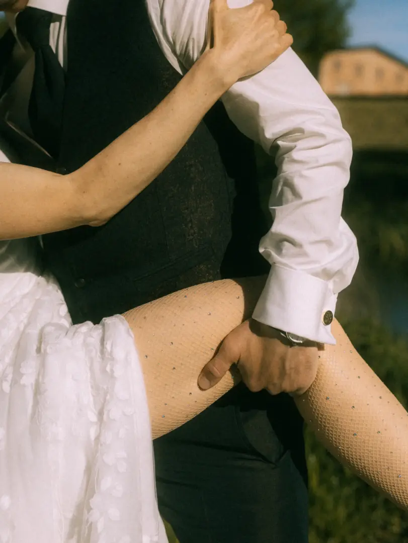 Intime Nahaufnahme eines Brautpaares bei Pärchenfotos in Schwerin: Der Bräutigam in Weste und weißem Hemd hält liebevoll das Bein seiner Braut, die eine mit Strass verzierte Netzstrumpfhose unter ihrem weißen Hochzeitskleid trägt. Eine romantische Szene im Freien bei warmem Sonnenlicht.