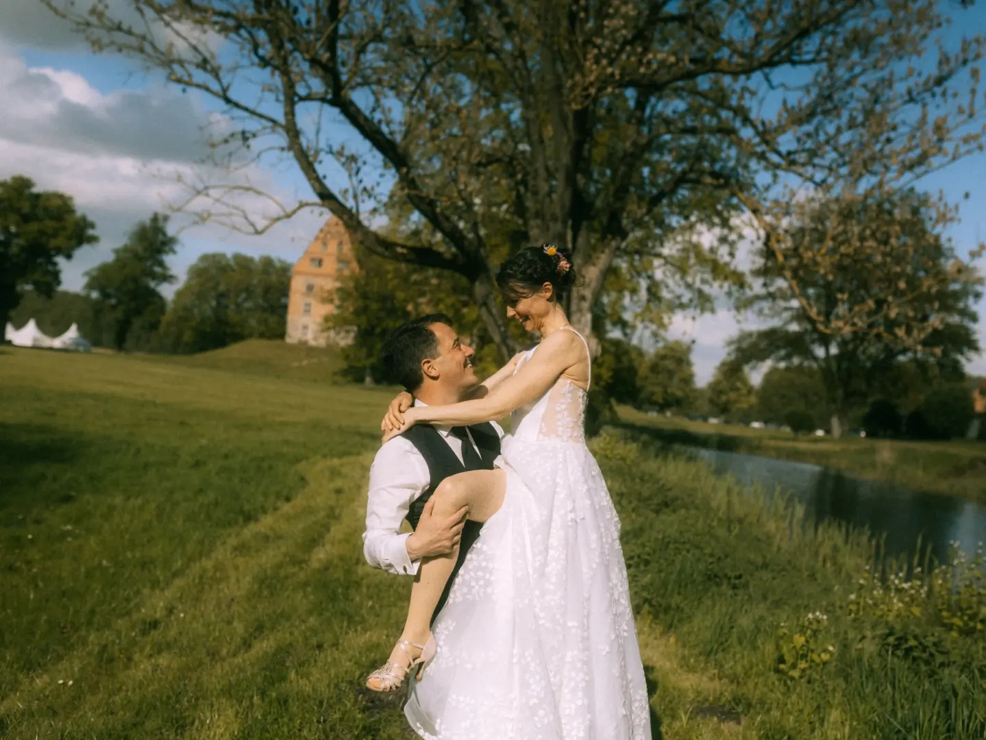 Ein frisch vermähltes Brautpaar in einer romantischen Pose: Der Bräutigam hebt seine Braut auf einer sonnigen Wiese vor einem historischen Schloss hoch. Die Braut trägt ein weißes Spitzenkleid und beide strahlen sich glücklich an. Lovestory Fotograf in Schwerin und MV.