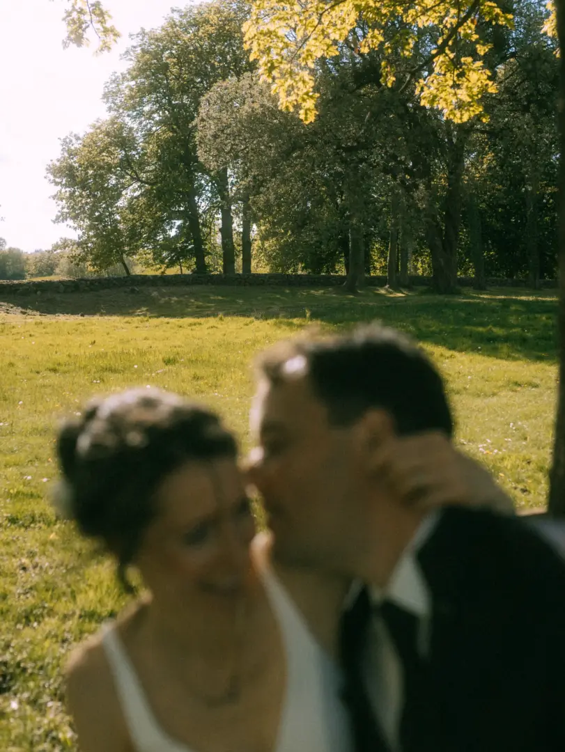 Ein unscharfes, romantisches Pärchenfoto im Freien, aufgenommen als Lovestory Fotograf in Schwerin und MV. Das Paar ist in eine liebevolle Umarmung vertieft, während im Hintergrund eine sonnendurchflutete Wiese mit alten Bäumen eine idyllische, warme Atmosphäre schafft.