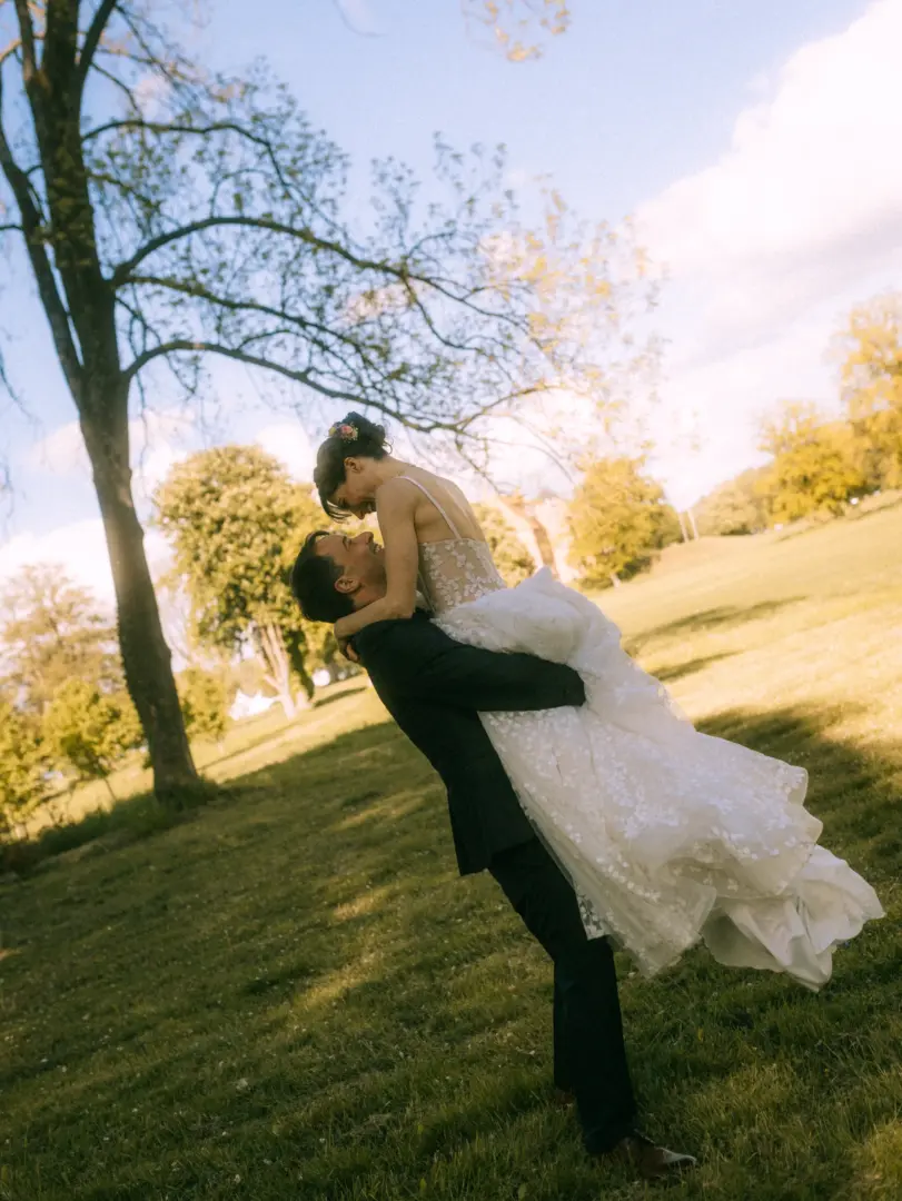 Ein glückliches Brautpaar in einer romantischen Pose: Der Bräutigam hebt seine Braut in einem weißen Spitzenkleid auf einer sonnigen Wiese hoch. Stimmungsvolle Aufnahme für Hochzeitsfotograf in Schwerin und MV, die die Freude des Paares in einer natürlichen Umgebung einfängt.