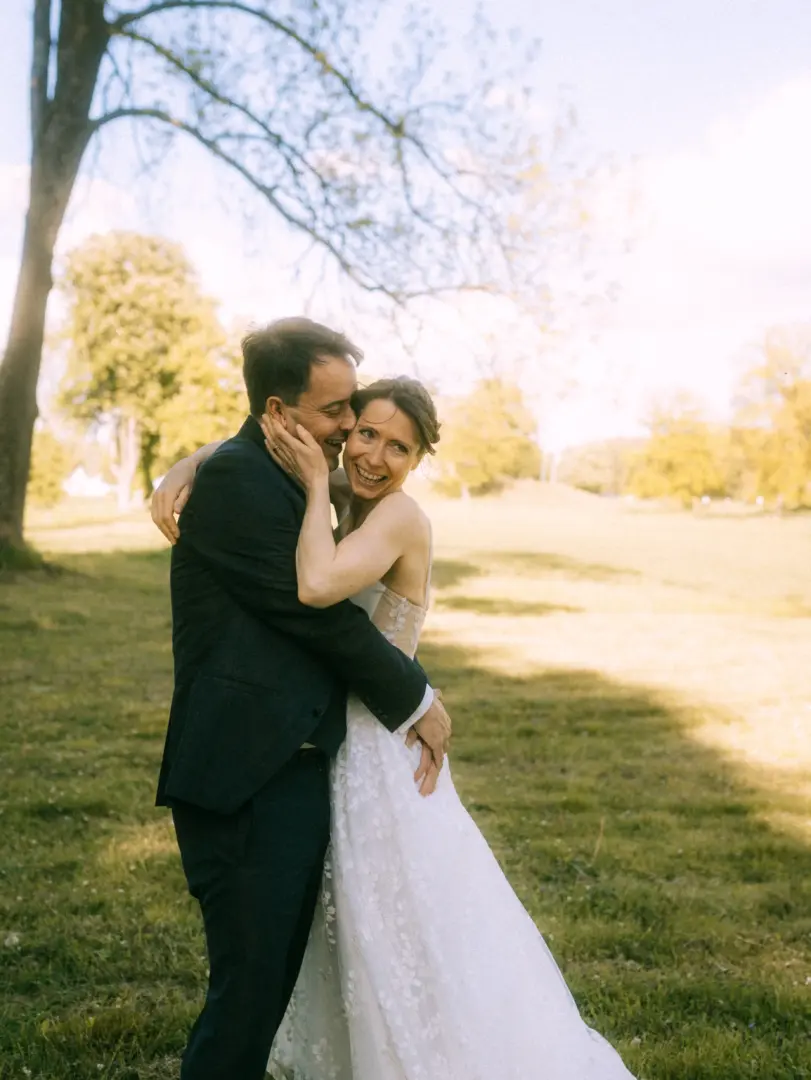 Ein glückliches Brautpaar umarmt sich herzlich auf einer sonnigen Wiese. Die Braut trägt ein elegantes, besticktes Hochzeitskleid und lacht, während der Bräutigam im dunklen Anzug sie zärtlich hält. Ein stimmungsvolles Bild vom Hochzeitsfotograf in Schwerin und MV.