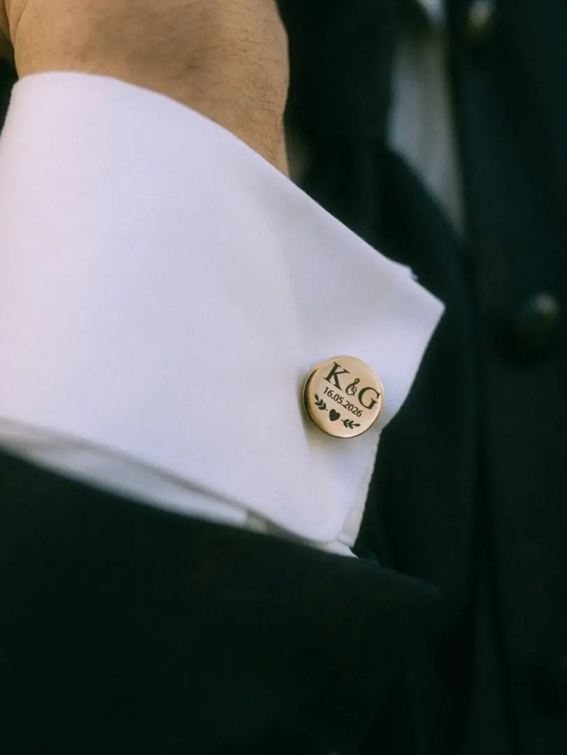 Nahaufnahme eines goldenen Manschettenknopfs an einem weißen Hemdärmel, graviert mit den Initialen „K&G“, dem Datum „16.05.2026“ sowie einem Herz- und Blättermotiv. Ein elegantes Detail für den Bräutigam, festgehalten von Ihrem Hochzeitsfotograf in Schwerin und MV.