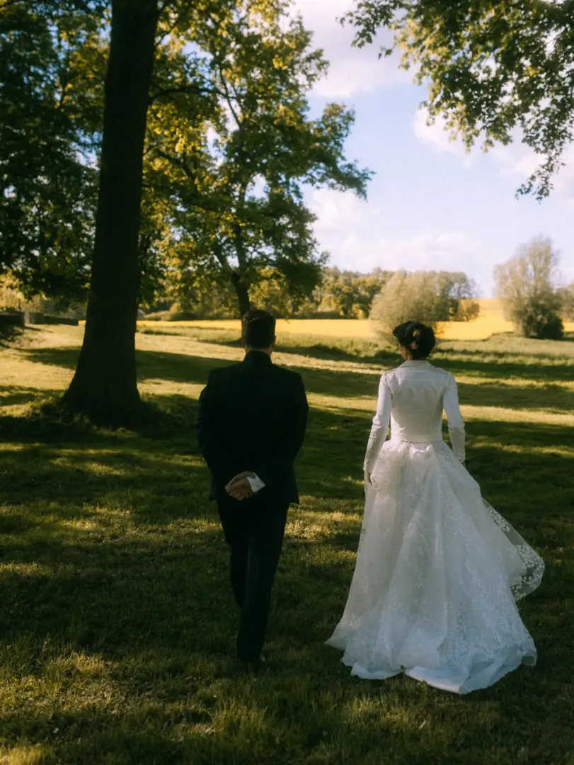 Ein Brautpaar geht von hinten gesehen Hand in Hand durch eine sonnige Wiese. Die Braut trägt ein elegantes, weißes Kleid mit Spitzenrock, während der Bräutigam einen dunklen Anzug trägt. Romantische Hochzeitsfotografie als Hochzeitsfotograf in Schwerin und MV.