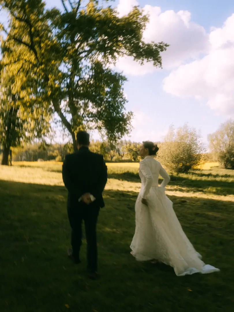 Ein Brautpaar spaziert bei goldenem Licht durch eine herbstliche Wiese unter einem großen Baum. Die Braut trägt ein elegantes weißes Spitzenkleid, während der Bräutigam im dunklen Anzug neben ihr geht. Eine stimmungsvolle Aufnahme von einem Hochzeitsfotograf in Schwerin und MV.