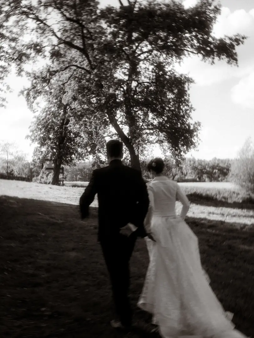 Ein Brautpaar rennt in einer verträumten, leicht unscharfen Schwarz-Weiß-Aufnahme über eine Wiese unter großen Bäumen. Die romantische Szene fängt die Dynamik ihrer Lovestory ein. Hochzeitsfotograf in Schwerin und MV.
