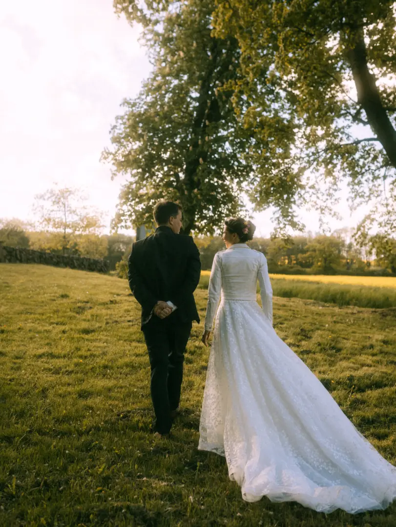 Ein Brautpaar geht Hand in Hand durch eine sonnendurchflutete Wiese bei Sonnenuntergang. Die Braut trägt ein weißes Spitzenkleid mit passender Jacke, der Bräutigam einen eleganten dunklen Anzug. Romantische Stimmung in der Natur, festgehalten von einem Hochzeitsfotograf in Schwerin und MV.
