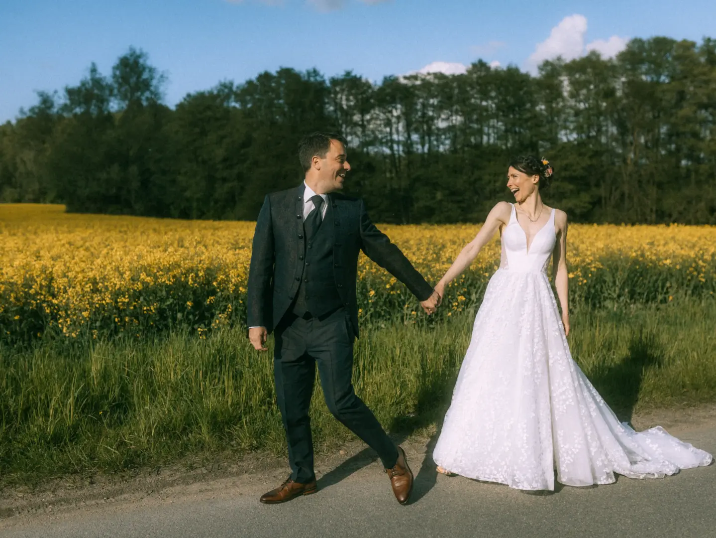 Ein glückliches Brautpaar hält sich an den Händen und lacht sich an, während sie gemeinsam an einem sonnigen Rapsfeld entlanglaufen. Die Braut trägt ein elegantes weißes Hochzeitskleid, der Bräutigam einen dunklen Anzug. Eine authentische Aufnahme vom Hochzeitsfotograf in Schwerin und MV.