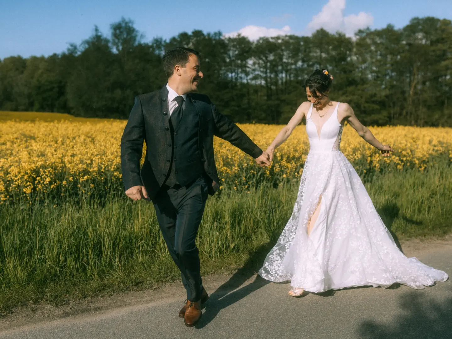 Ein glückliches Brautpaar läuft lachend an der Hand an einem leuchtend gelben Rapsfeld entlang. Die Braut trägt ein elegantes, weißes Spitzenkleid mit Beinschlitz, der Bräutigam einen dunklen Anzug. Eine authentische Aufnahme vom Hochzeitsfotograf in Schwerin und MV.