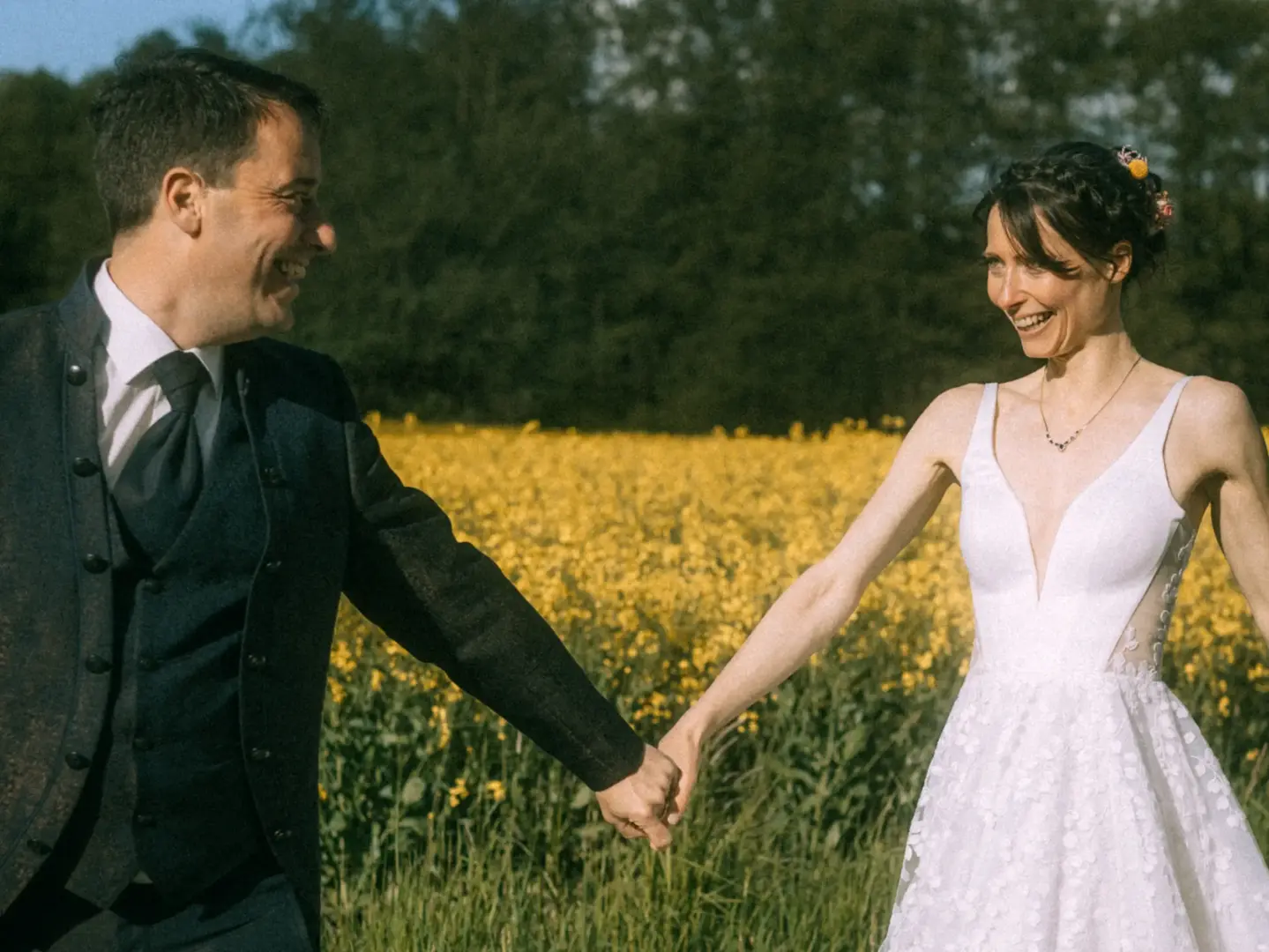 Ein glückliches Brautpaar hält sich in einem sonnigen Rapsfeld an den Händen und lacht sich verliebt an. Die Braut trägt ein weißes Spitzenkleid, der Bräutigam einen eleganten dunklen Anzug. Eine stimmungsvolle Aufnahme von einem Lovestory Fotograf in Schwerin und MV.