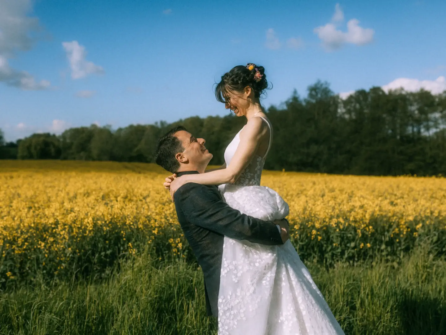 Ein glückliches Brautpaar in einem sonnigen Rapsfeld: Der Bräutigam hebt seine Braut im weißen Spitzenkleid liebevoll in die Höhe, während beide sich anlächeln. Eine romantische Szene für ein Paarshooting, festgehalten von Ihrem Lovestory Fotograf in Schwerin und MV.