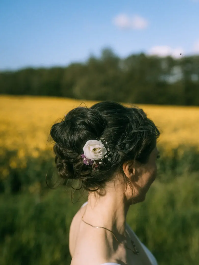 Eine Braut mit kunstvoll hochgestecktem Haar und einer eleganten Blumenverzierung aus einer weißen Rose und kleinen lila Akzenten blickt vor einem sonnigen, leuchtend gelben Rapsfeld in die Ferne – eingefangen von Ihrem Hochzeitsfotograf in Schwerin und MV.