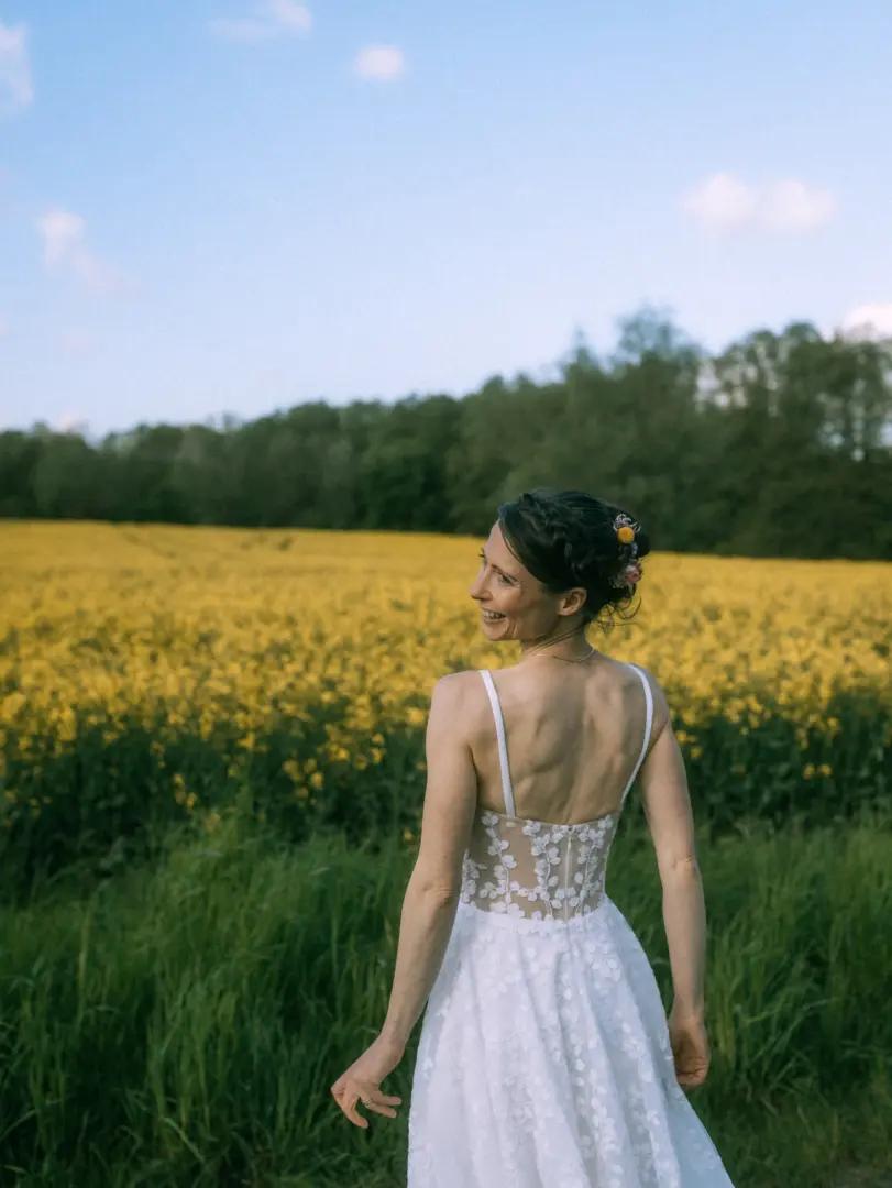Eine Braut mit geflochtener Hochsteckfrisur und Blumenschmuck blickt lächelnd über die Schulter, während sie in einem weißen Spitzenkleid vor einem leuchtend gelben Rapsfeld steht. Eine stimmungsvolle Aufnahme vom Hochzeitsfotograf in Schwerin und MV.