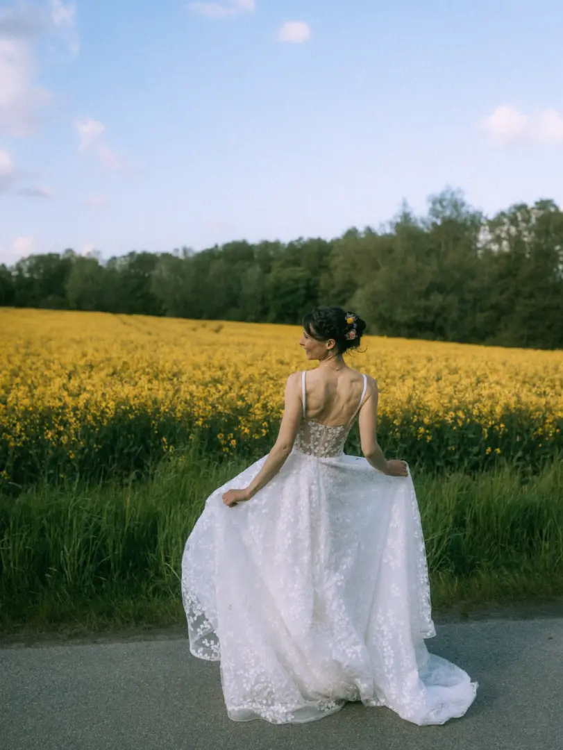 Eine Braut in einem eleganten weißen Spitzenkleid blickt lächelnd über die Schulter, während sie durch ein sonniges Rapsfeld spaziert. Die romantische Szene im Freien wurde von einem Hochzeitsfotograf in Schwerin und MV festgehalten.