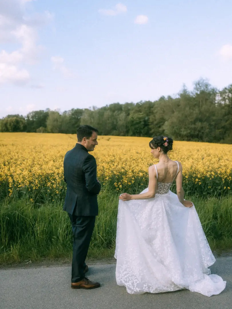 Ein Brautpaar blickt sich liebevoll auf einer Landstraße vor einem leuchtend gelben Rapsfeld an. Die Braut trägt ein detailliertes Spitzenkleid, der Bräutigam einen eleganten dunklen Anzug. Eine romantische Szene, eingefangen von einem Lovestory Fotograf in Schwerin und MV.