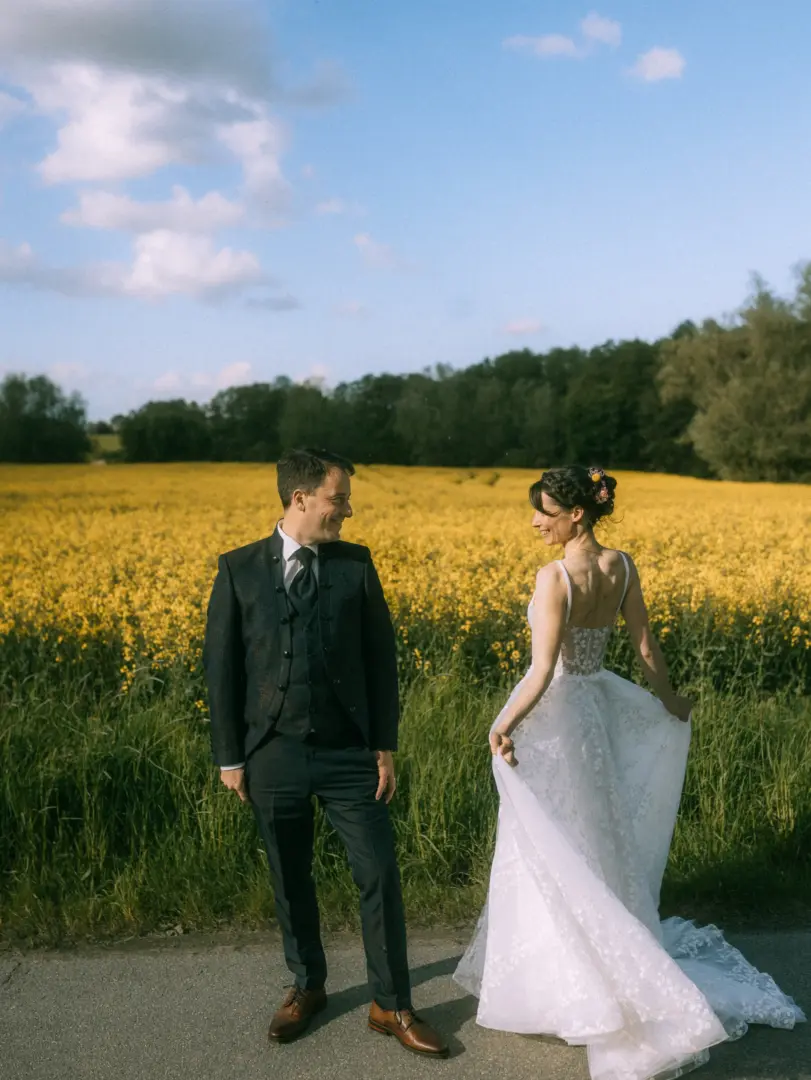 Ein glückliches Brautpaar blickt sich in einem sonnigen Rapsfeld liebevoll an. Die Braut trägt ein elegantes Spitzenkleid und hält es leicht in den Händen, während der Bräutigam einen dunklen Anzug trägt. Eine romantische Szene als Lovestory Fotograf in Schwerin und MV festgehalten.