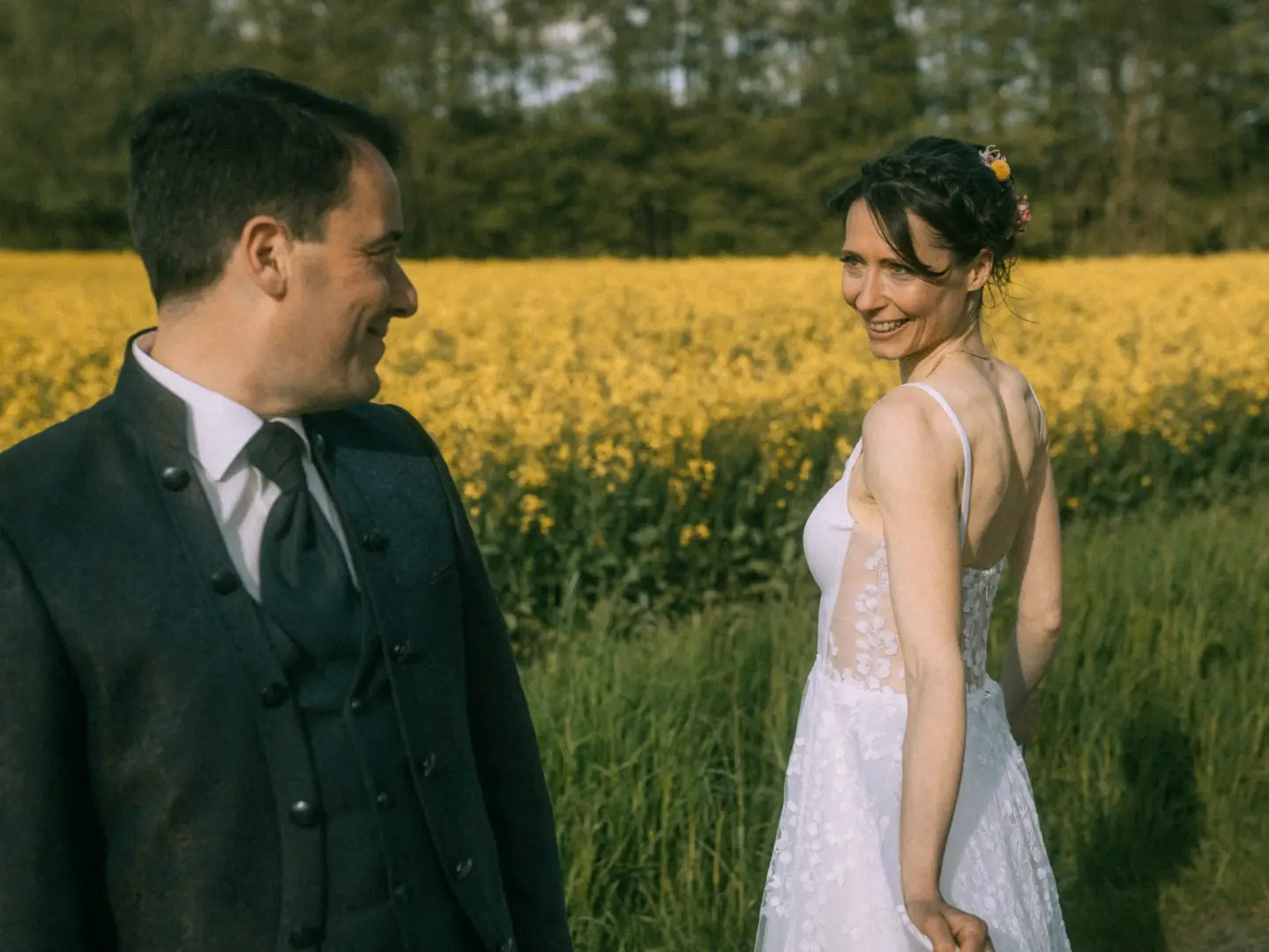 Ein glückliches Brautpaar blickt sich in einem blühenden Rapsfeld liebevoll an. Die Braut trägt ein weißes Spitzenkleid mit Rückenausschnitt und Blumen im Haar, der Bräutigam einen eleganten Anzug. Eine romantische Szene für eine Lovestory als Fotograf für Pärchen in Schwerin.