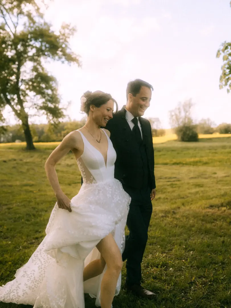 Ein glückliches Brautpaar spaziert lachend über eine sonnige Wiese. Die Braut trägt ein elegantes, weißes Spitzenkleid und hält es leicht hoch, während der Bräutigam im dunklen Anzug neben ihr geht. Eine authentische Aufnahme vom Lovestory Fotograf in Schwerin und MV.